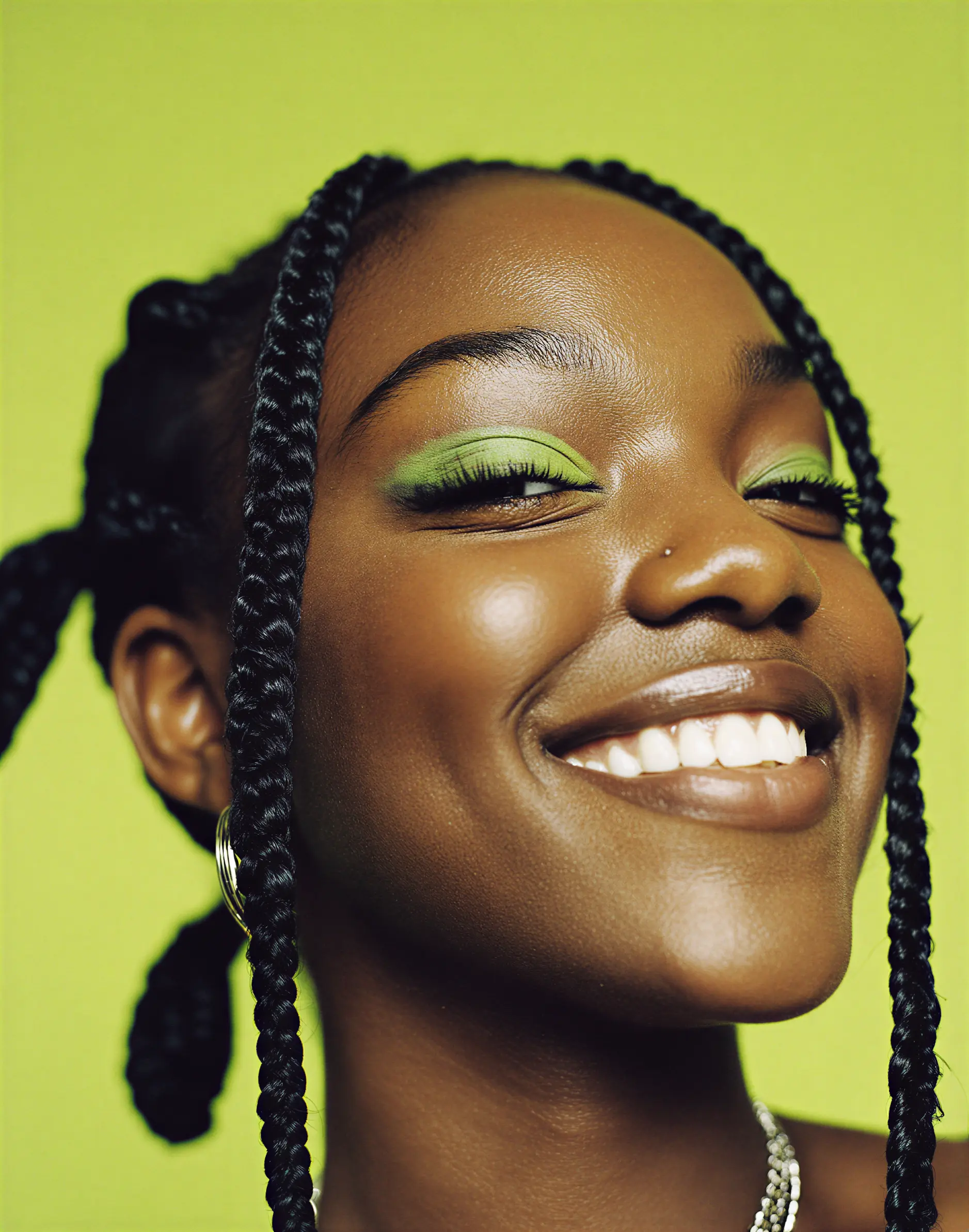 Close-up portrait of a smiling woman with braided hair and green eyeshadow against a green background.