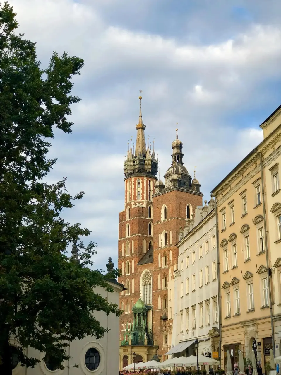 St. Mary's Basilica with its two distinctive towers rising behind historic buildings and a leafy tree under a partly cloudy sky.