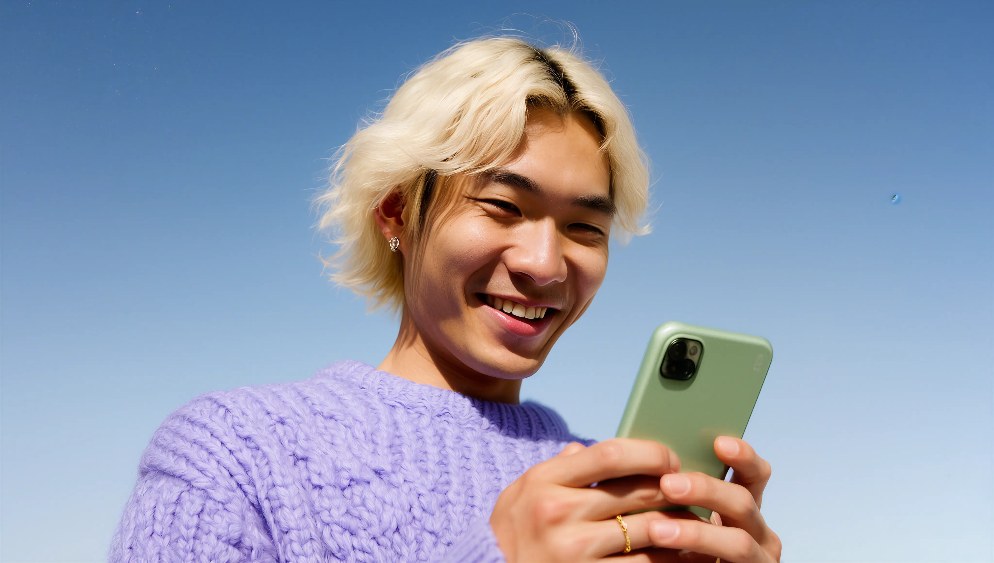 Smiling young person with blonde hair wearing a purple sweater looking at a green smartphone outdoors against a clear blue sky.